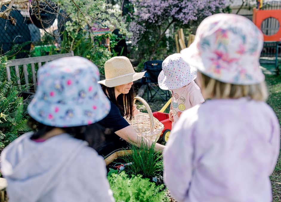 Two female children, wearing hats, watch an educator and another child working in a vegetable garden at Canning Vale Prekindy. 