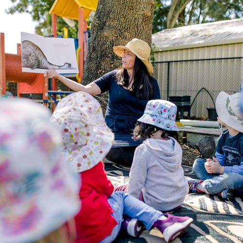 An educator holds up a book with an emu illustration while she reads to four children in the foreground. The educator and children are outside in a playground. 