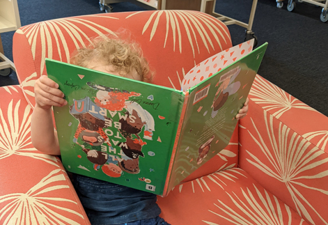 A child sits in an orange chair holding a book upside down. 