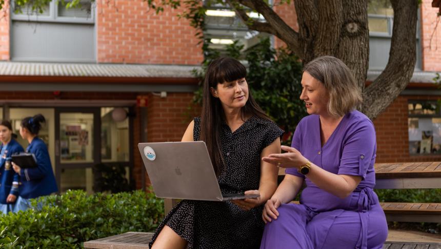 Two female teachers have a conversation while viewing a laptop in the school yard.