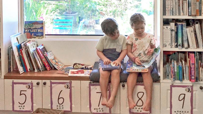 A boy and a girl sit on a window seat looking at a book. Shelves of books surround them on the left and right.
