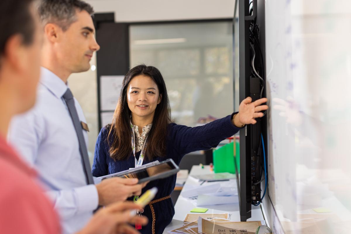 Three teachers stand close to a whiteboard in a classroom. A young, Asian teacher points to the board while the two other male teachers take notes. 