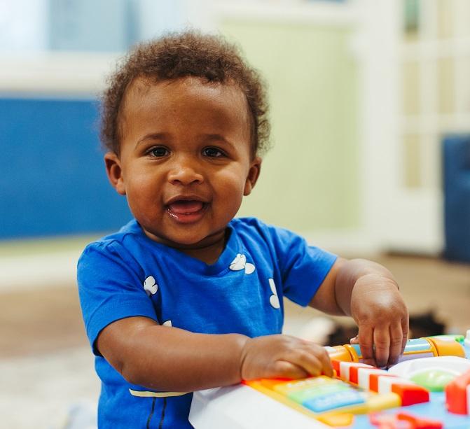 A small boy in a blue shirt in an early learning setting, looking at the camera and with his hands on a colourful toy. 